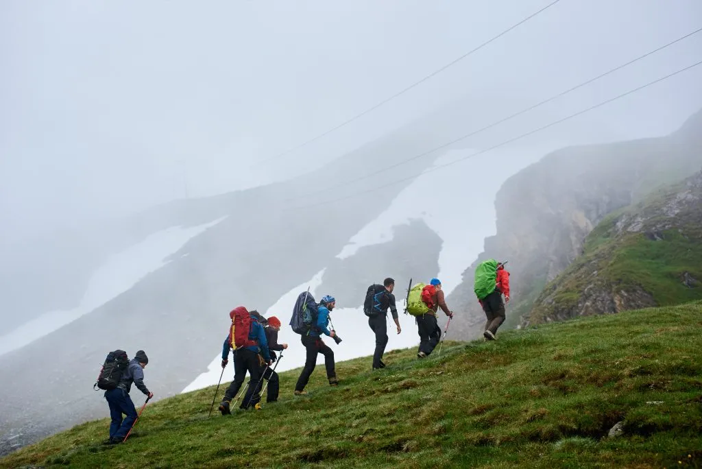 Back view of male hikers with backpacks climbing grassy hill. Young tourists walking uphill in mountains with foggy cliff on background. Concept of hiking, travelling and backpacking.